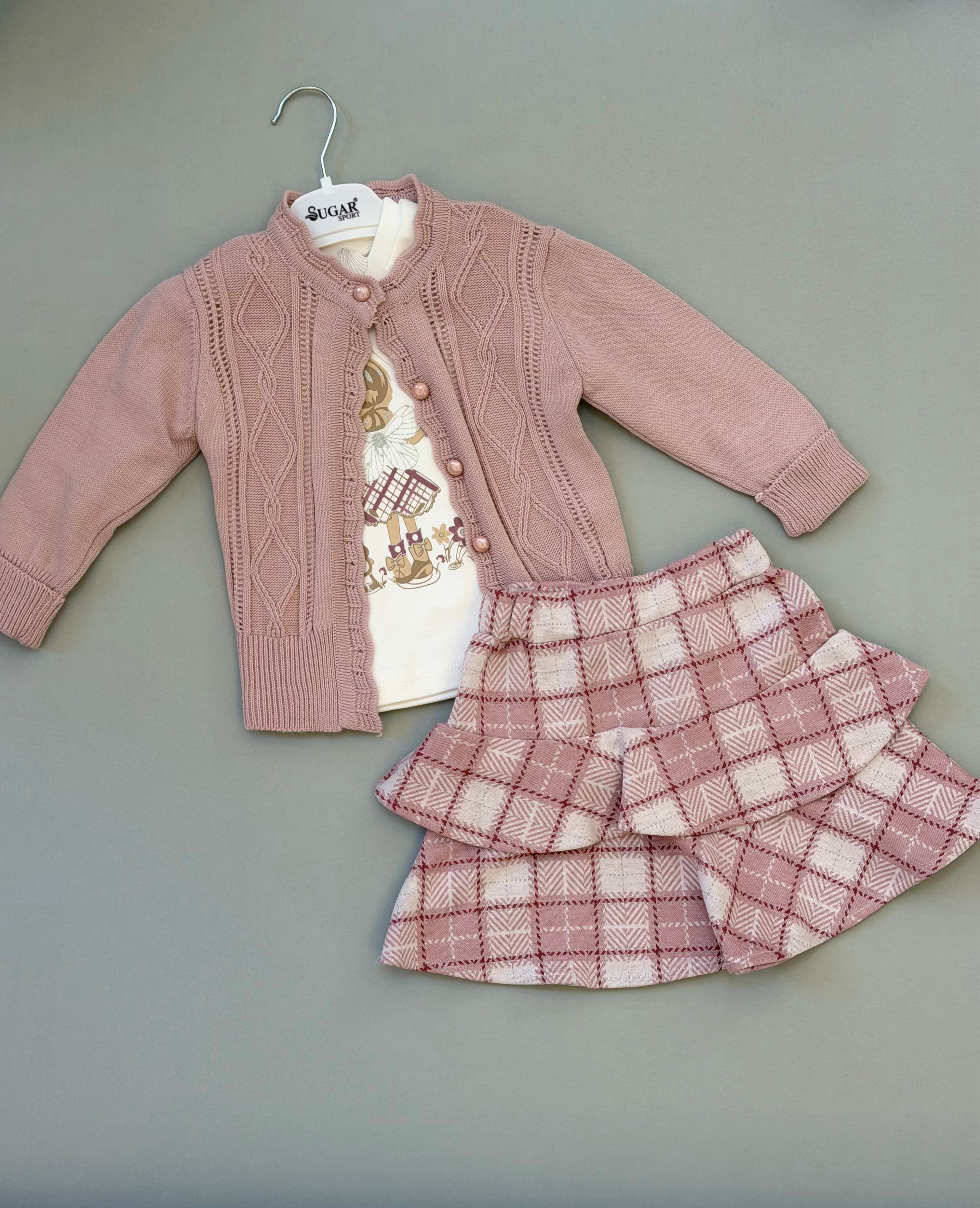 Children's outfit with pink cardigan, white shirt, and plaid skirt on a hanger against a gray background